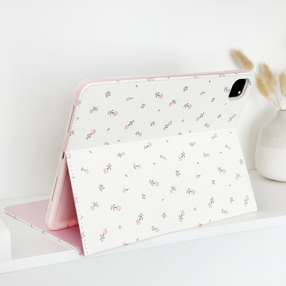 A tablet with the Coconut Lane Ditsy Floral White iPad Case, made from vegan leather and featuring a light pink and white ditsy floral pattern, is displayed on a white shelf. Next to it, there is a small white vase holding some dried grass stalks, adding a touch of nature to the minimalist setup. The anti-slip feature ensures it stays securely in place.