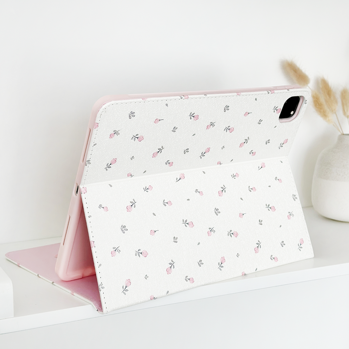 A tablet with the Coconut Lane Ditsy Floral White iPad Case, made from vegan leather and featuring a light pink and white ditsy floral pattern, is displayed on a white shelf. Next to it, there is a small white vase holding some dried grass stalks, adding a touch of nature to the minimalist setup. The anti-slip feature ensures it stays securely in place.