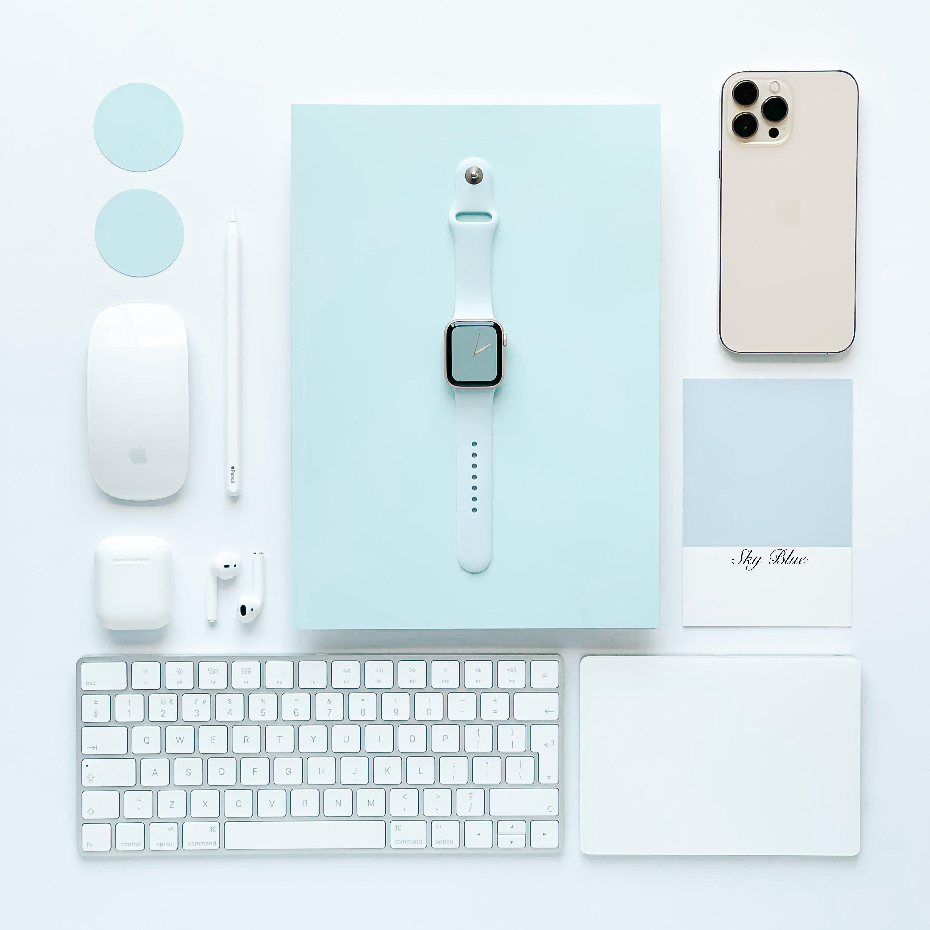 A neatly arranged flat lay featuring tech accessories in a minimalistic design. Includes an Apple Watch with a Coconut Lane NAKD Apple Watch Strap - Sky Blue, iPhone, keyboard, mouse, stylus, AirPods, two circular discs, and a card reading 