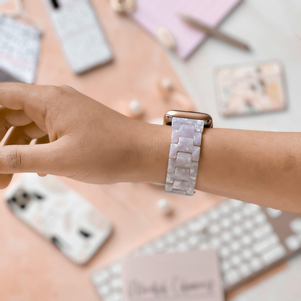 A hand is shown wearing the Coconut Lane Luxe Pearl Apple Watch Strap, featuring a light purple, marbled design and an adjustable strap. In the background, there are smartphones in cases, a tablet, and a keyboard on a light pink surface. The overall scene suggests a stylish and tech-friendly workspace.