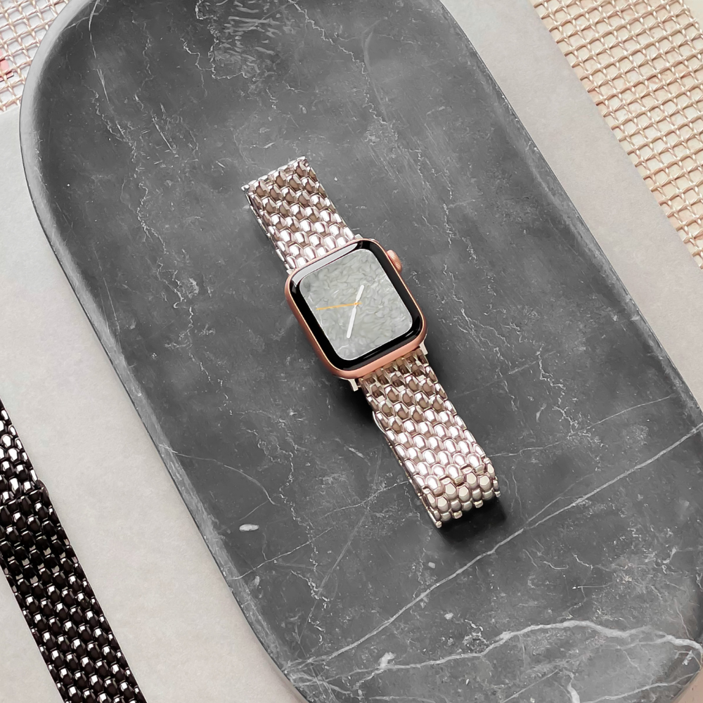 A smartwatch featuring a Mermaid Watch Strap - Silver from Coconut Lane is placed on an oval, dark grey marble tray. The watch face displays an analog clock with gold hands. The tray rests on a flat surface, surrounded by various materials in different colors and textures.