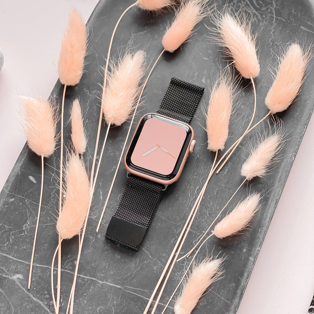 A Coconut Lane Magnetic Chain Apple Watch Strap in black, featuring a sleek pink-faced watch, is elegantly positioned on a dark marbled tray. Surrounding the watch are several artistically arranged dried, fluffy, light beige flowers. The overall setting exudes a stylish and minimalist vibe.